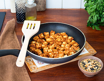 Gebratene Tofuwürfel in einer Pfanne mit Dipp auf einem Holzbrett mit Dekoration.