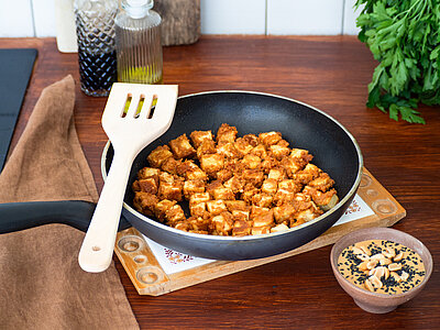 Gebratene Tofuwürfel in einer Pfanne mit Dipp auf einem Holzbrett mit Dekoration.