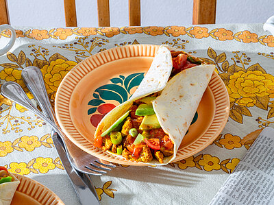 Ein Burrito mit Tofu und buntem Gemüse auf einem Teller, der auf einem Tischtuch steht.