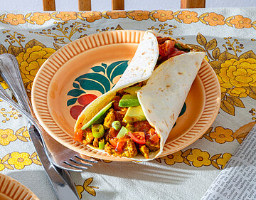 Breakvast-Burrito Ein Burrito mit Tofu und buntem Gemüse auf einem Teller, der auf einem Tischtuch steht.