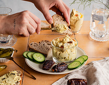 Hellbraune Creme mit Datteln, Gurkenscheiben und Brotscheiben auf einem Teller mit Tischdekoration.