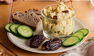 Hellbraune Creme mit Datteln, Gurkenscheiben und Brotscheiben auf einem Teller mit Tischdekoration.