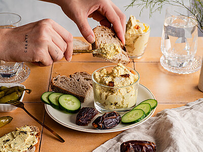 Hellbraune Creme mit Datteln, Gurkenscheiben und Brotscheiben auf einem Teller mit Tischdekoration.