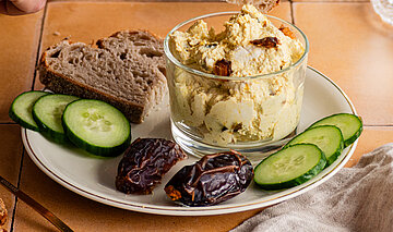 Hellbraune Creme mit Datteln, Gurkenscheiben und Brotscheiben auf einem Teller mit Tischdekoration.
