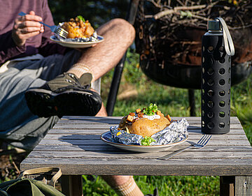 Aufgeschnittene Backkartoffel mit Tofu-Würfeln, Salat und Joghurt auf einem Teller neben einer Wasserflasche auf einem Holztisch.