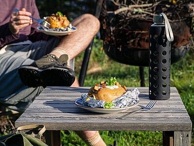 Aufgeschnittene Backkartoffel mit Tofu-Würfeln, Salat und Joghurt auf einem Teller neben einer Wasserflasche auf einem Holztisch.