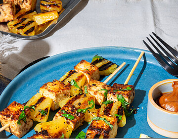 Drei Tofu-Spiesse mit Dip und Früchtedekoration auf blau-weißem Tischtuch.