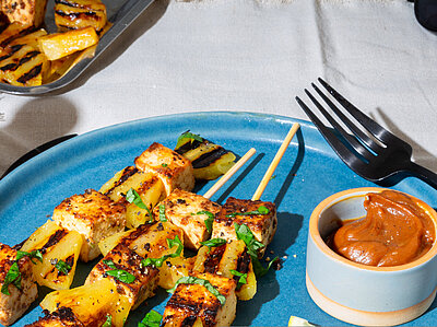 Drei Tofu-Spiesse mit Dip und Früchtedekoration auf blau-weißem Tischtuch.