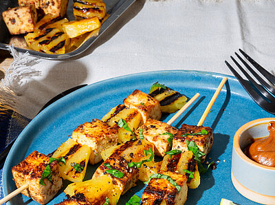 Drei Tofu-Spiesse mit Dip und Früchtedekoration auf blau-weißem Tischtuch.