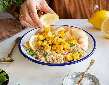 Gelbe Tofuwürfel auf Couscous und Fladenbrot auf einem Teller, der von einer Hand gehalten wird, mit Tischdekoration.