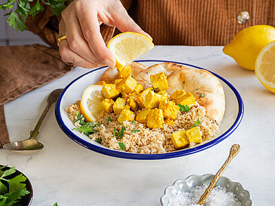 Gelbe Tofuwürfel auf Couscous und Fladenbrot auf einem Teller, der von einer Hand gehalten wird, mit Tischdekoration.