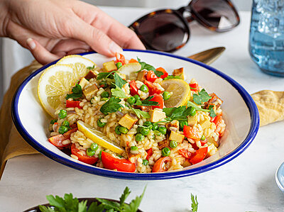 Paella mit Tofustückchen und buntem Gemüse und Zitrone auf einem Teller mit Tischdekoration.