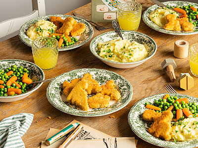 Mehrere Teller mit Tofu Nuggets, Kartoffelpüree und Erbsen und Möhren mit Tischdekoration.