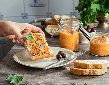 Roter Aufstrich auf einem Stück Ciabatta mit Oliven, daneben ein Glas mit dem Aufstrich und Dekoration.