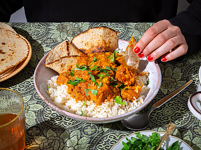 Tofuwürfel mit roter Soße auf Reis mit dünnem Fladenbrot auf einem Teller mit Tischdekoration. Eine Hand am Teller.