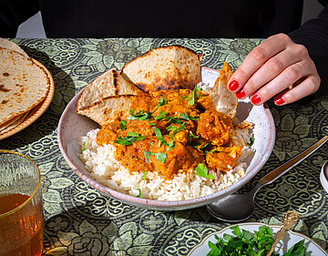 Tikka Masala Tofuwürfel mit roter Soße auf Reis mit dünnem Fladenbrot auf einem Teller mit Tischdekoration. Eine Hand am Teller.