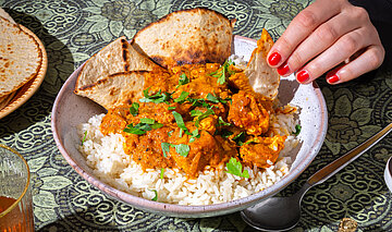 Tofuwürfel mit roter Soße auf Reis mit dünnem Fladenbrot auf einem Teller mit Tischdekoration. Eine Hand am Teller.