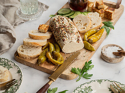 Teilweise aufgeschnittenes Brot auf einem Holzbrett mit heller Creme, Oliven und Peperoni mit Tischdekoration.