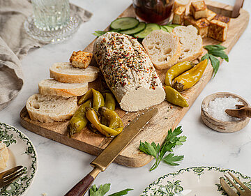Teilweise aufgeschnittenes Brot auf einem Holzbrett mit heller Creme, Oliven und Peperoni mit Tischdekoration.