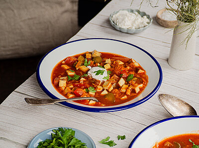 Tofuwürfel in einer roten Suppe mit Gemüse und einem Klecks weißer Creme auf 2 Tellern mit Tischdekoration.