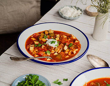 Tofuwürfel in einer roten Suppe mit Gemüse und einem Klecks weißer Creme auf 2 Tellern mit Tischdekoration.
