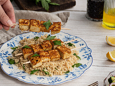Tofu-Spiesse auf Couscous auf einem Teller mit buntem Gemüse und Tischdekoration.