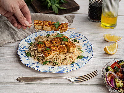 Tofu-Spiesse auf Couscous auf einem Teller mit buntem Gemüse und Tischdekoration.