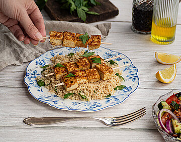 Tofu-Spiesse auf Couscous auf einem Teller mit buntem Gemüse und Tischdekoration.