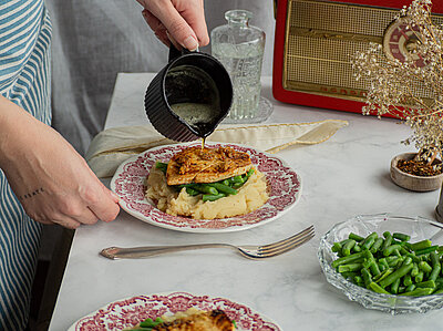 Tofu auf Kartoffelpüree mit Bohnen auf Teller, flüssige Butter wird auf die Tofuscheibe gegossen.