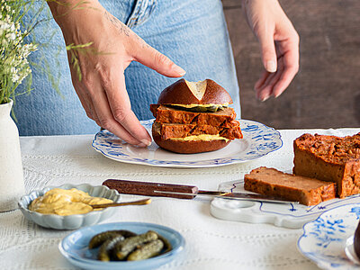 2 Hände halten ein Brötchen mit 2 Leberkäs-Scheiben mit Senf auf einem Teller mit Tischdekoration.