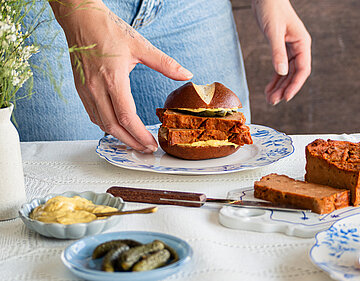 2 Hände halten ein Brötchen mit 2 Leberkäs-Scheiben mit Senf auf einem Teller mit Tischdekoration.