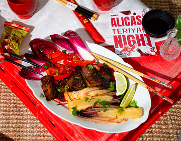 Dunkle Tofuscheiben an Gemüse auf einem Teller mit Tischdekoration. Jemand hält Essstäbchen.