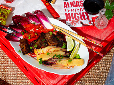 Dunkle Tofuscheiben an Gemüse auf einem Teller mit Tischdekoration. Jemand hält Essstäbchen.