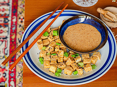 Helle Tofustückchen mit Lauch auf einem Teller mit Dip und Tischdekoration.
