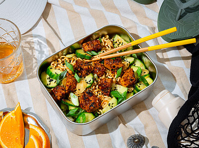 Nudelsalat mit Gurken und gewürztem Tofu in einer Metallschüssel auf einem Tischtuch mit Deko.