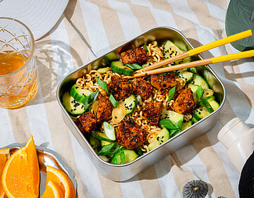 Nudelsalat mit Gurken und gewürztem Tofu in einer Metallschüssel auf einem Tischtuch mit Deko.