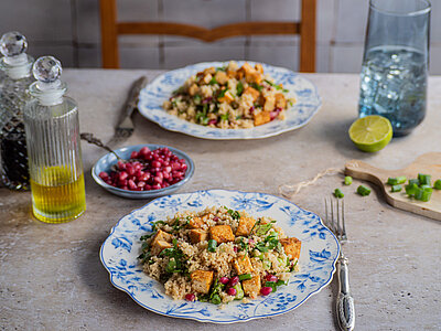 Couscous mit Tofustückchen Lauch, Petersiele und Grantapfelkernen auf 2 Tellern mit Tischdekoration.