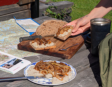 Helle Creme mit Zwiebeln und Tofustückchen auf einem Teller und Broten, die von einer Hand gehalten werden mit Tischdekoration.