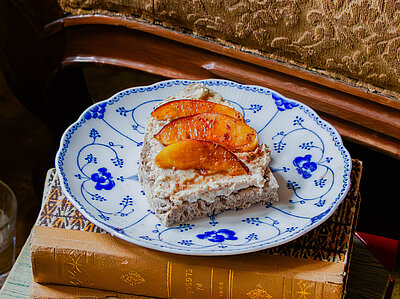 Eine Scheibe Brot mit Tofu-Aufstrich und 3 Pfirsichscheiben auf einem Bücherstapel vor einem Sof, auf dem ein Buch liegt.