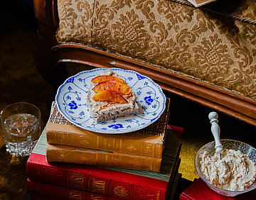 Eine Scheibe Brot mit Tofu-Aufstrich und 3 Pfirsichscheiben auf einem Bücherstapel vor einem Sof, auf dem ein Buch liegt.