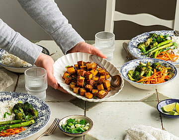 Einige Teller mit Räuchertofu und Gemüse auf einem Tisch, ein Teller wird in die Kamera gehalten. 