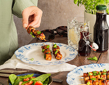 Zwei Grillspieße auf einem Teller, ein weiterer wird in die Kamera gehalten, mit Dekoration.