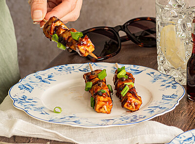 Zwei Grillspieße auf einem Teller, ein weiterer wird in die Kamera gehalten, mit Dekoration.