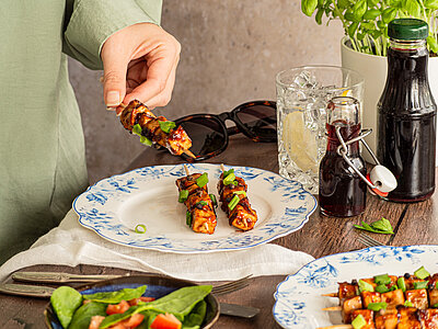 Zwei Grillspieße auf einem Teller, ein weiterer wird in die Kamera gehalten, mit Dekoration.