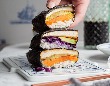 Eine Hand hält Sushi in 3 Algenblättern mit Tofu, Reis, Rotkohl und Süßkartoffel auf einem Brett mit Tischdekoration.