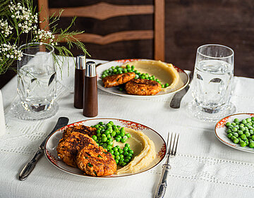 Tofu-Frikadellen mit Kartoffelpüree und Erbsen auf 2 Tellern mit Tischdekoration.