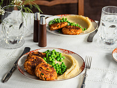 Tofu-Frikadellen mit Kartoffelpüree und Erbsen auf 2 Tellern mit Tischdekoration.