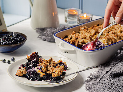 Tofu-Crumble mit Heidelbeeren in einer Schüssel und einem Teller mit Tischdekoration.