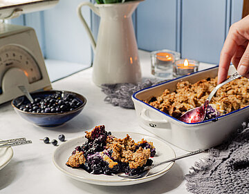 Tofu-Crumble mit Heidelbeeren in einer Schüssel und einem Teller mit Tischdekoration.