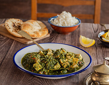 Palak Paneer Tofuwürfel mit Spinat-Soße auf einem Teller mit kleinen Schüsseln auf einem Holztisch.